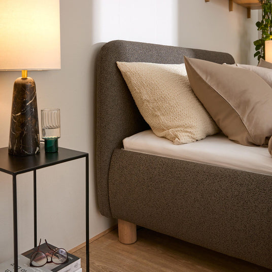 Närbild av bäddad säng med beige kuddar, stickad pläd och smalt nattygsbord med mörk marmorlampa, glas och böcker i ett varmt och ombonat sovrum.