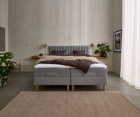 Minimalistiskt sovrum med grå stoppad dubbelsäng, neutralt bäddset i bruna och beige toner, växter, vägglampor, en inramad affisch och ljusa gardiner.