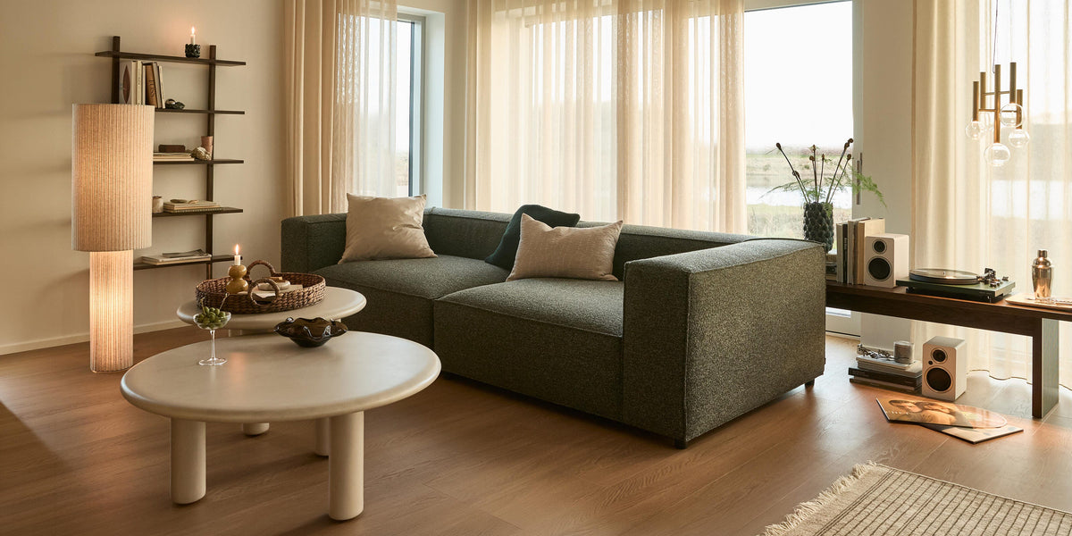 Ljus, skandinavisk vardagsrum med grå modulsoffa, runda ljusa soffbord, skira beige gardiner, tända ljus och golvlampa, hylla med böcker samt konsol med skivspelare och högtalare; matta och växt vid stora fönster i dagsljus.