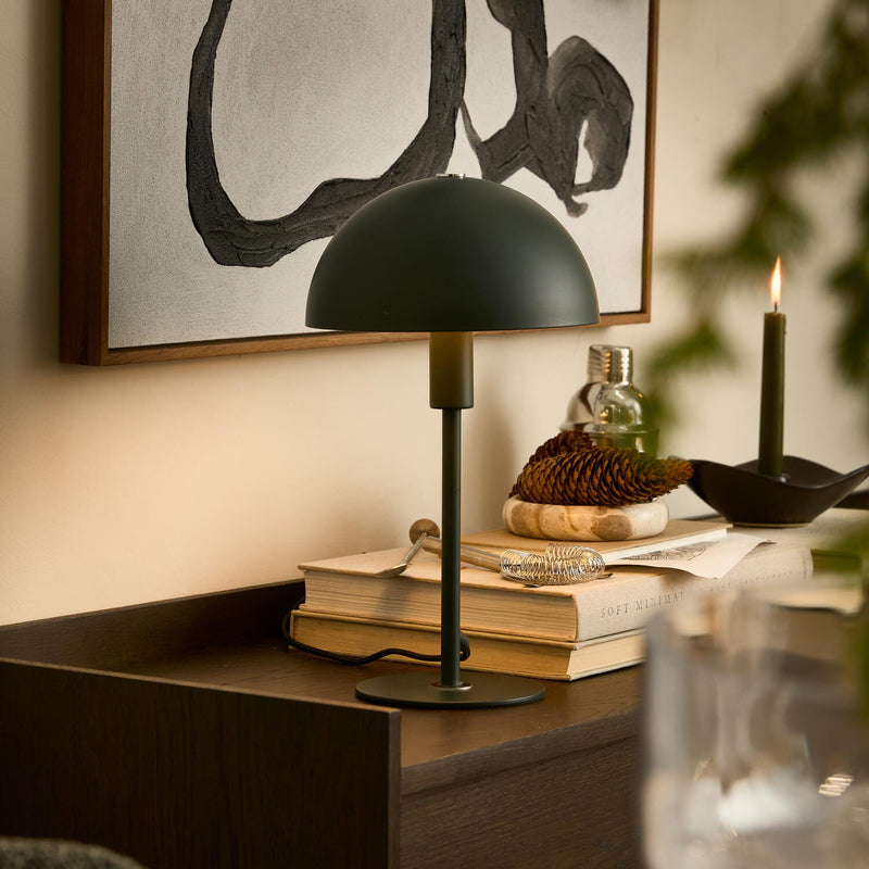 Mörkgrön bordslampa på böcker med "SOFT MINIMAL", tallkotte och tändt ljus på ett sidobord i en varm, modern interiör med abstrakt väggkonst.