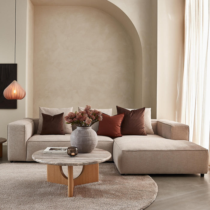 Beige vardagsrum med hörnsoffa, runt soffbord i sten och trä, bruna kuddar, vas med blommor och hängande lampa i mjukt dagsljus.