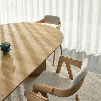 Ljust matbord i trä med två stoppade stolar, gröna glasskålar och skira vita gardiner i dagsljus.