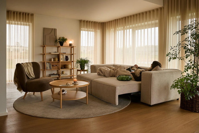 Ljust, varmt vardagsrum i nordisk stil med beige modulsoffa, rund matta och träbord; skira beige gardiner, gröna växter och ett tänt ljus medan en person vilar under en filt.