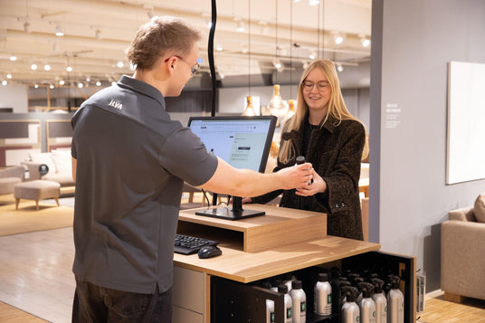 Butiksmedarbetare i ILVA-tröja räcker över en liten flaska till kund vid disk med skärm som visar ILVAs webbplats i ett möbelshowroom.