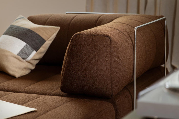 Närbild på brun modulsoffa med metallarmstöd och kudde i beige och grått vid ett fönster med ljusa gardiner.