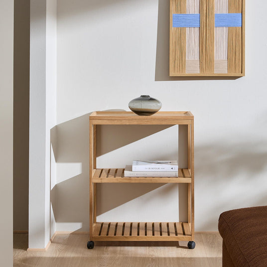 Minimalistisk hylla i ljus ek på små hjul med ribbade hyllplan; liten keramikvas ovanpå, tavla med blå detaljer på väggen och böckerna Soft Minimal och The Kinfolk Home på mittenhyllan i ett ljust vardagsrum med solljus.