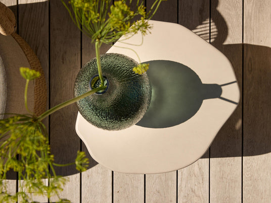 Grön texturerad glasvas med blommor på ett ljust organiskt sidobord på trädäck i solsken, sedd ovanifrån.