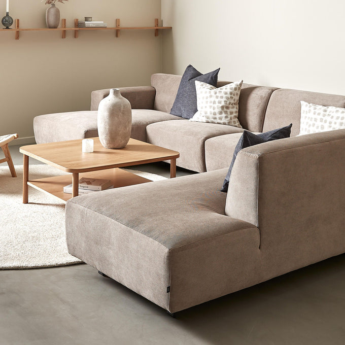 Beige hörnsoffa med träsoffbord, neutrala kuddar och vägg­hylla med vas och ljus i ett ljust vardagsrum.