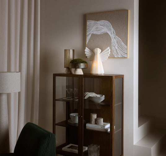 Vitrinskåp i mörkt trä med glasdörrar, lampängel och vas med mossa ovanpå, abstrakt tavla på väggen, boken “Kinfolk” på hyllan samt skålar, glas och ljus; beige gardiner och dämpad, varm belysning.