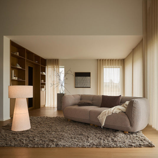 Ljust vardagsrum med beige gardiner, stor brunbeige soffa på mjuk ryamatta, golvlampa och inbyggda hyllor vid stora fönster i dagsljus.