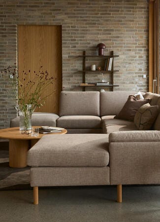 Vardagsrum med L-formad beige soffa, träbord, golvlampa och glasvas med kvistar; stora fönster och tegelvägg.