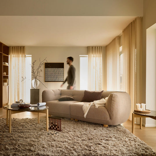Lugn beige vardagsrum med rundad soffa, mjuk ryamatta, runda soffbord, golvlampa och skira gardiner; en person går förbi.