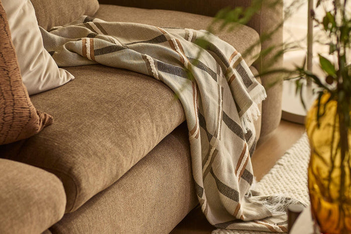 Närbild på beige/brun soffa med randig pläd och kuddar; ravfärgad vas och ljus, strukturerad matta i ett varmt vardagsrum.