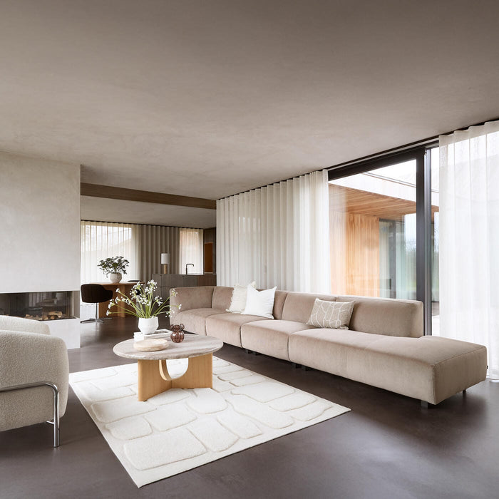 Ljust, minimalistiskt vardagsrum med beige modulsoffa, vit strukturerad matta, trä-sofabord med vas och blommor, stora skjutdörrar i glas och skira gardiner.