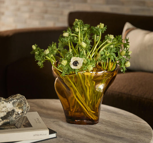 Amberfärgad glasvas med gröna stjälkar och vita anemoner på runt marmorbord; bredvid ligger en sten och två böcker, varav en med titeln HOTEL Weekend.