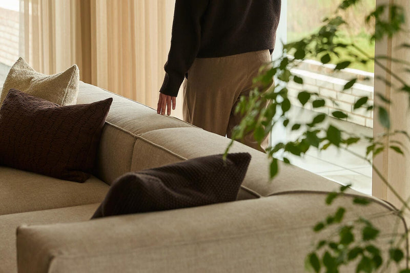 Beige soffa med bruna och cremefärgade kuddar intill skira gardiner; en person står vid fönstret med gröna växter i förgrunden.
