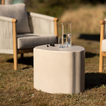 Beige utomhusbord i stenlook med karaff och glas med vatten mellan två loungestolar på gräsmatta i sol.