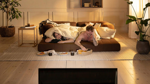 Par ligger under täcke på låg soffmodul och ser på TV med snacks och läsk i ett varmt, solbelyst vardagsrum med stora växter, matta och gardiner.