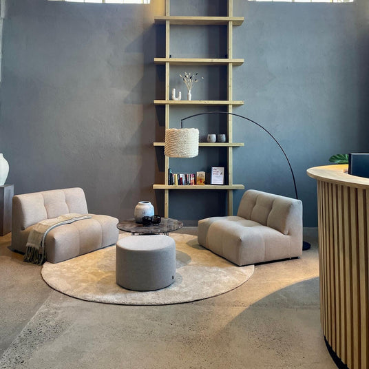 Loungehörna med två beige fåtöljer, rund matta, puf och soffbord framför vägg med hyllsystem, bågformad golvlampa och stora fönster.