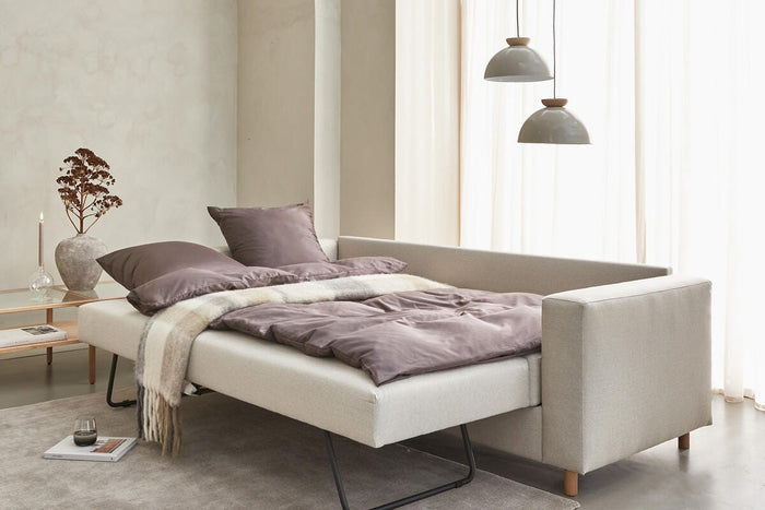 Beige bäddsoffa utdragen med mauvefärgat bäddset, två hängande pendlar, skira gardiner och sidobord med vas och ljus i ljus skandinavisk vardagsrumsmiljö.
