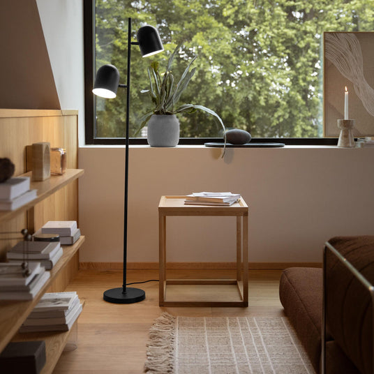 Minimalist hörn i vardagsrum med svart golvlampa med två skärmar, öppna trähyllor med böcker, litet kubformat sidobord i ek och en ljus matta; på fönsterkarmen står en krukväxt, ett ljus och en tavla, med dagsljus och grönska utanför.