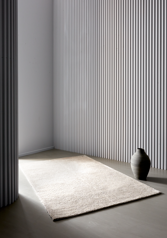 Ljus beige matta i en minimalistisk studio med räfflad vit vägg och grå vas i mjukt dagsljus.