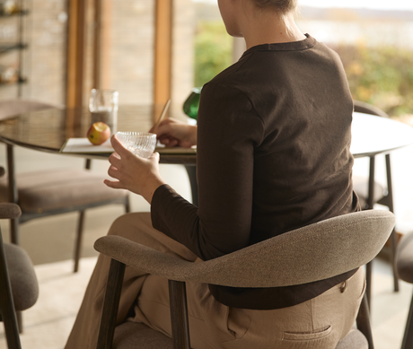 Person sitter på en stoppad spisestol vid ett runt matbord, håller ett glas och antecknar i ett ljust, luftigt matrum.