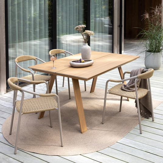 Uteplats med matbord i trä och fyra flätade stolar på trädäck, vas med blommor och skål på bordet, pläd över en stol och krukväxt i bakgrunden i dagsljus.