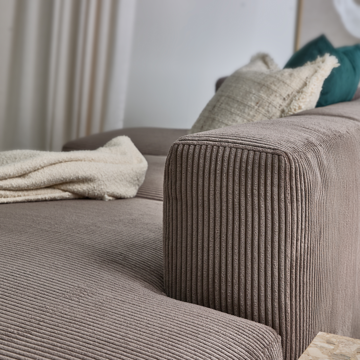 Närbild på en ribbad taupefärgad soffa med armstöd, en ljus cremefärgad pläd och kuddar i beige och petrolgrönt.