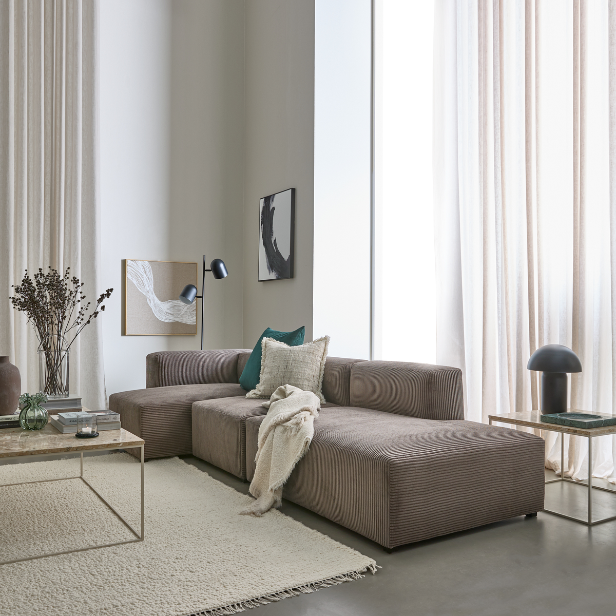 Ljus, skandinaviskt vardagsrum med beige modulsoffa, kuddar och pläd, glasbord, matta och höga skira gardiner i dagsljus.