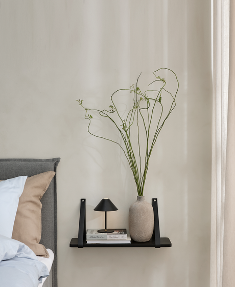 Väggmonterad hylla vid sängen med liten svart bordslampa, naturfärgad vas med långa slingrande kvistar och en bok med titeln “A Modern Way to Live” bredvid en grå sänggavel.