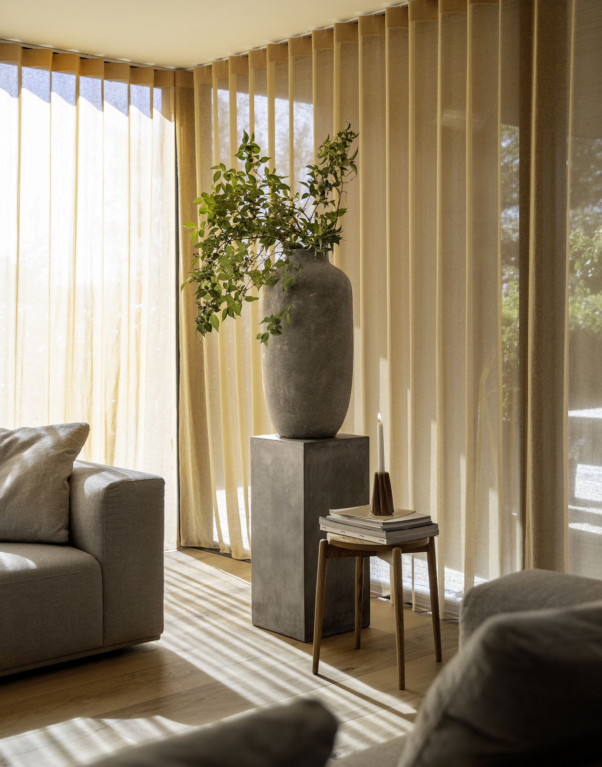 Solbelyst vardagsrum med golvlånga beige gardiner, grå soffa, stenvase med gröna kvistar på piedestal och liten träpall med böcker och ett ljus.