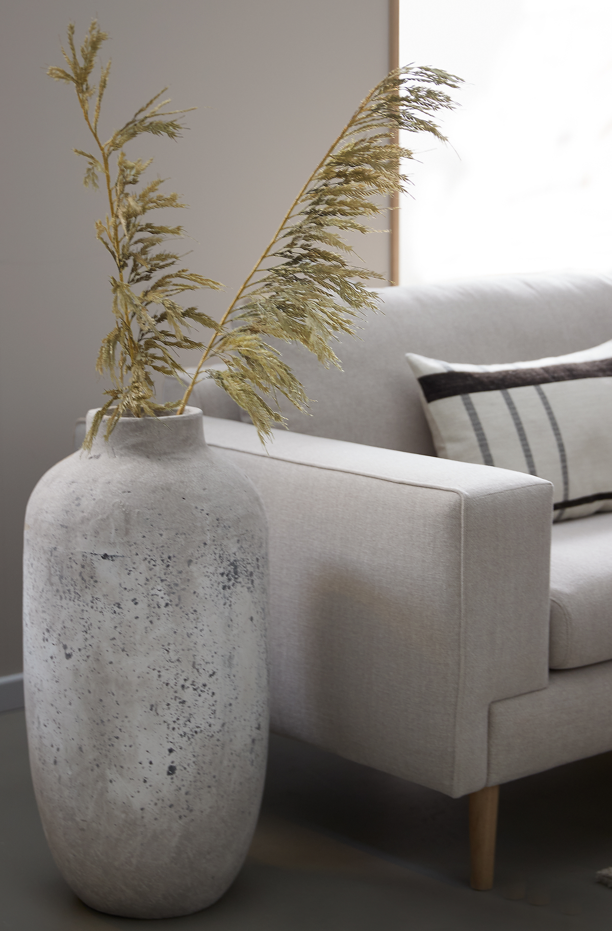 Stor betonglik vas med torkade strån bredvid en ljus beige soffa med kudde i ett ljust vardagsrum.