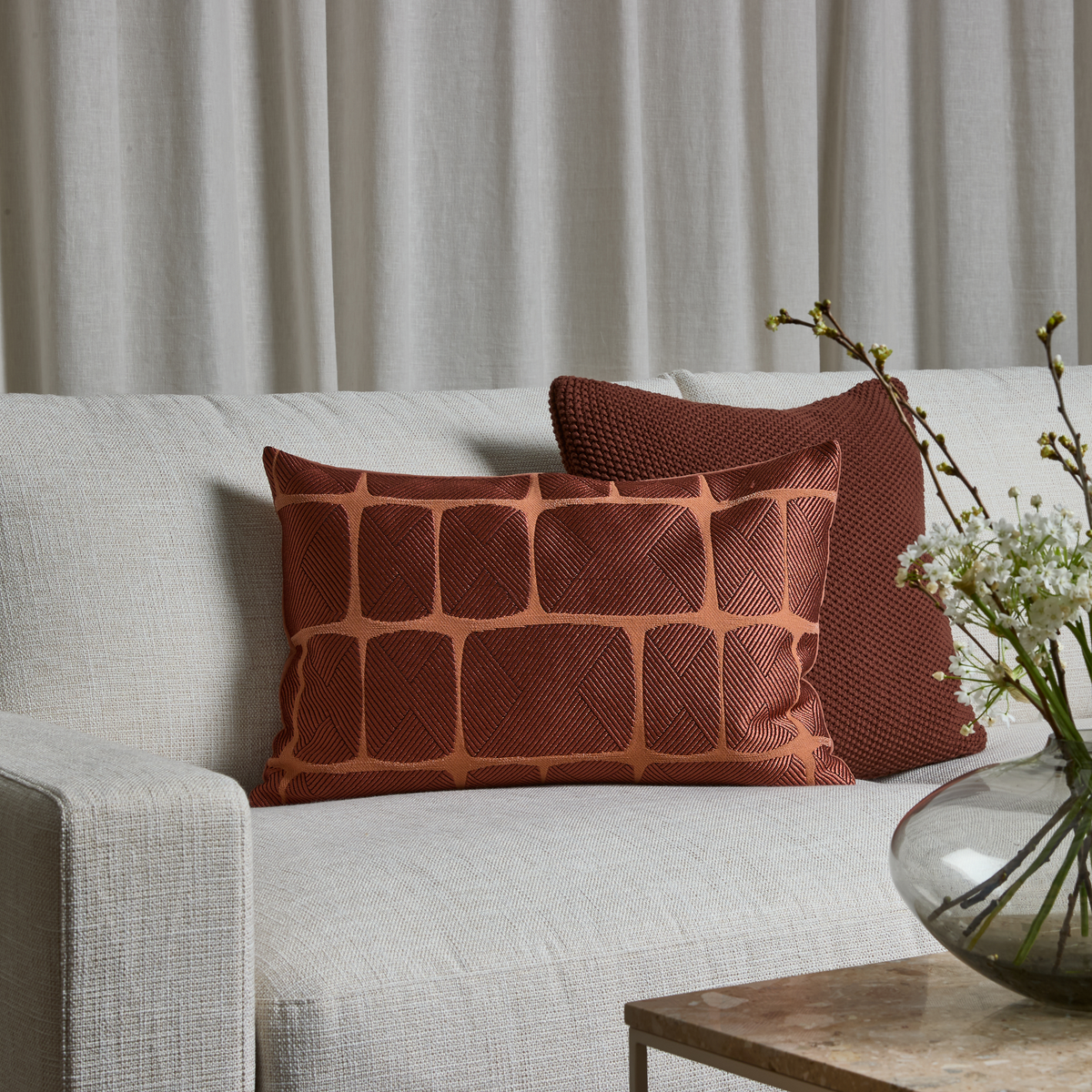 Ljus beige soffa med två rostfärgade prydnadskuddar, en avlång med geometriskt mönster, framför ljusa gardiner; glasvas med vita blommor på bord i förgrunden.