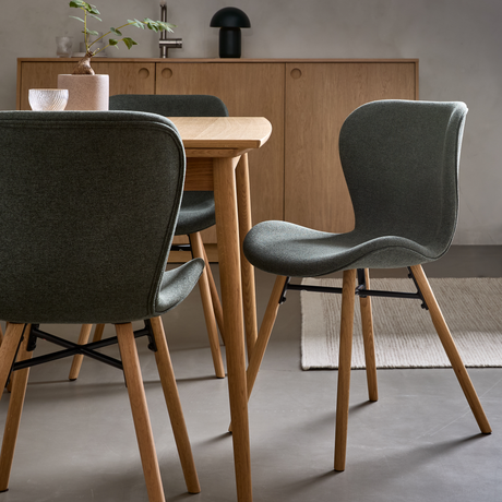 Matplats med träbord, grågröna stoppade stolar och sideboard i ek, med liten bordslampa och vas med kvist.