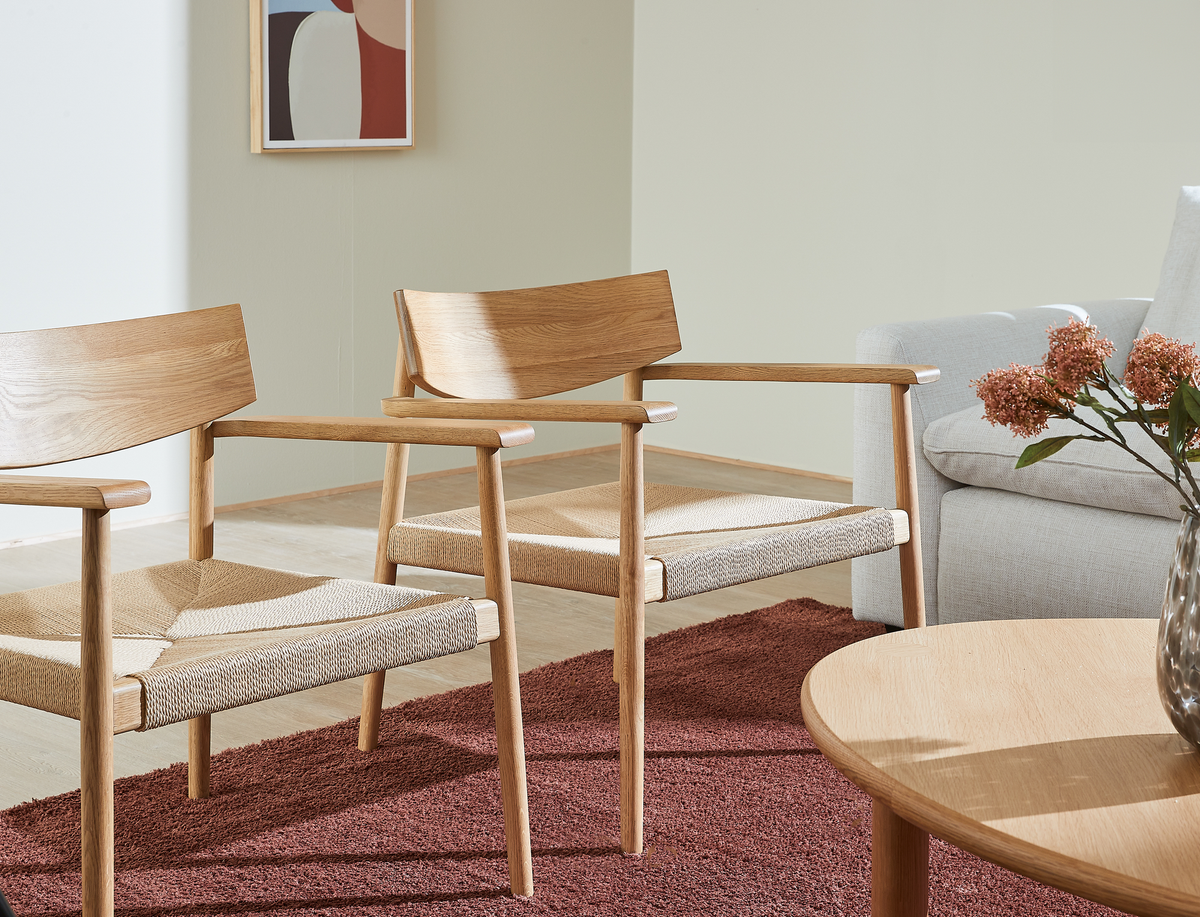 Två trästolar med vävd sits på rödbrun matta bredvid en ljusgrå soffa, rund träbordsskiva och vas med rosa blommor; abstrakt tavla på väggen.