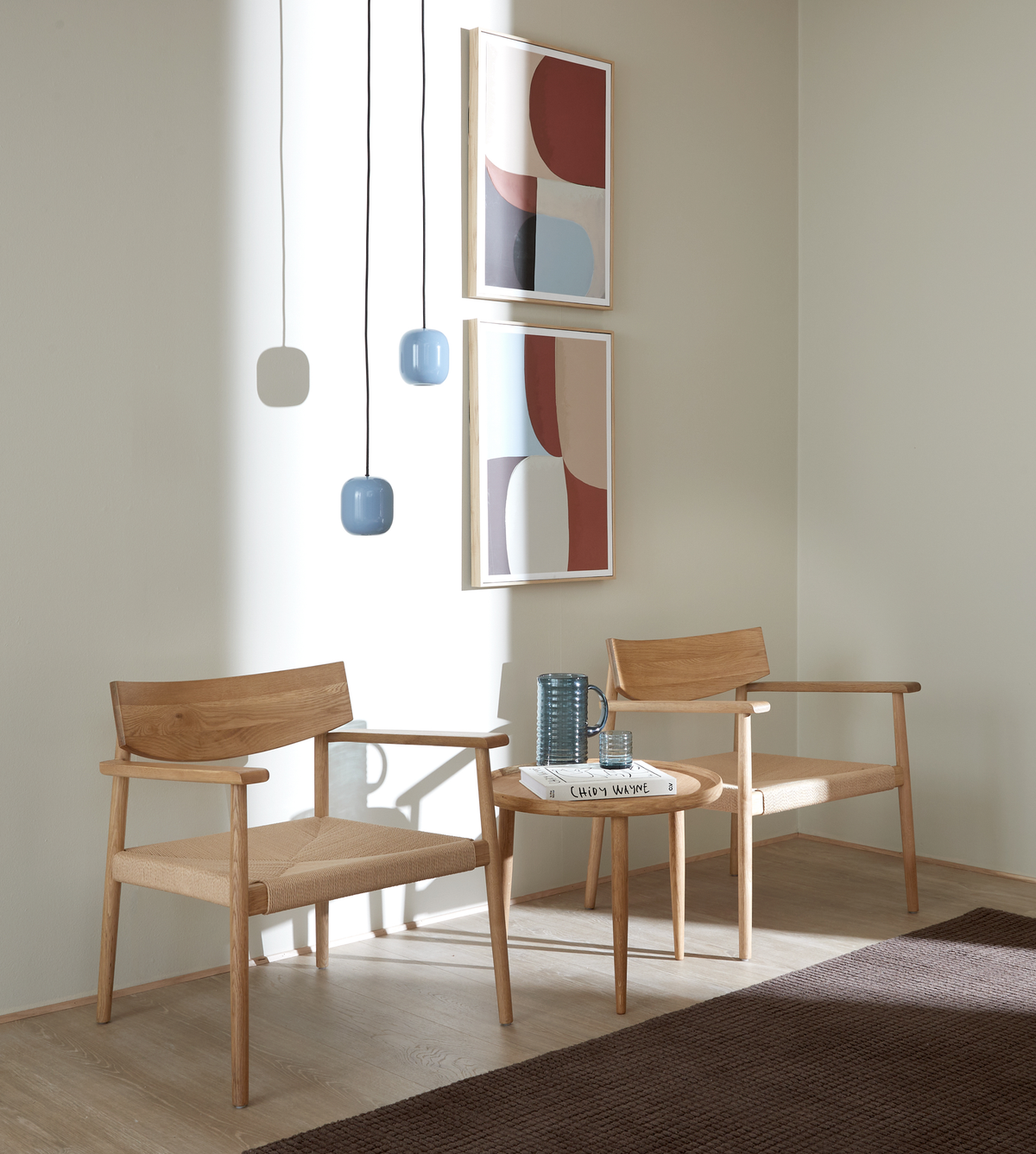 Två träfåtöljer med naturfärgade sitsar kring ett litet runt bord med en blå kanna, ett glas och boken ”CHLO WANE”; två abstrakta tavlor och tre blå pendellampor i mjukt dagsljus.