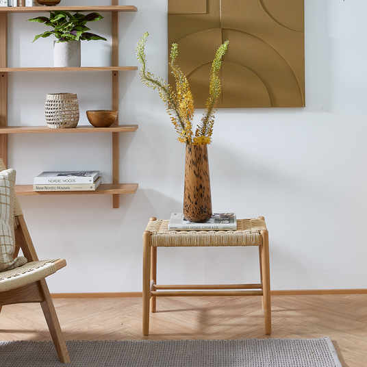 Ljus skandinavisk vardagsrumshörna: flätad pall med två böcker, ”THE HOME UPGRADE” och ett magasin, toppad med brunprickig vas med gula kvistar; vägghyllor med växt, korg och böcker ”NORDIC MOODS” och ”New Nordic Houses” i neutrala trä- och beige toner.