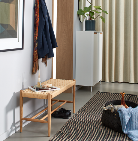Hall med vävd träbänk, hängd kappa och sjal, bricka med doftstickor, vitt skåp med krukväxt, beige gardiner och mönstrad matta; korg med leksaker och blå filt.