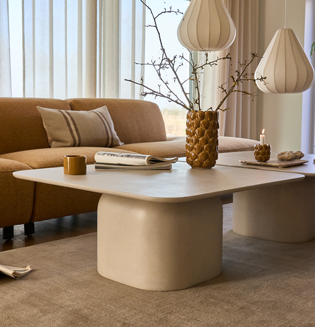Ljus vardagsrumsscen med beige soffbord med rundade hörn, senapsgul soffa, vita pendellampor, keramikvas med grenar, tändt ljus, mugg och tidning på bordet.