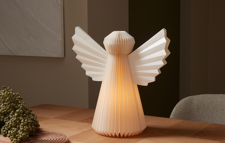 Vikbar papperslampa formad som en ängel med varmt sken på ett träbord i ett mysigt rum.