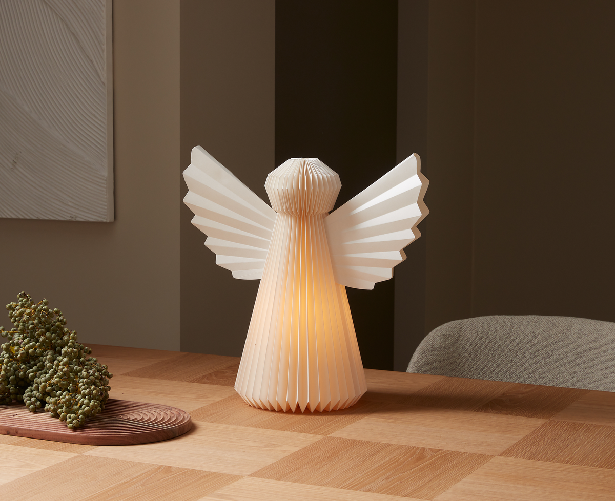 Vikad pappersängel‑formad bordslampa som lyser varmt på ett träbord i en minimalistisk interiör.