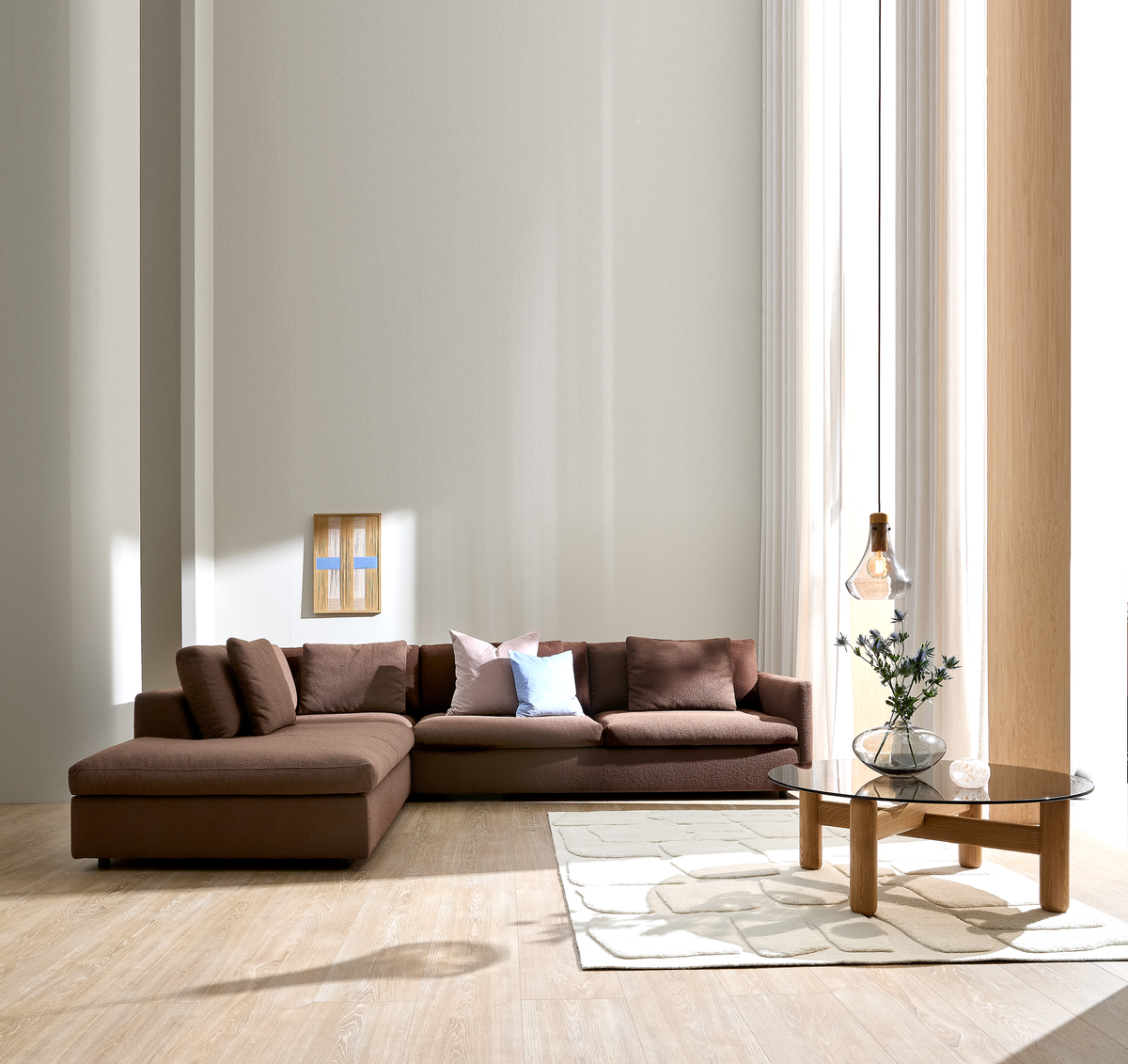 Ljus, minimalistisk vardagsrumsmiljö med brun modulsoffa, ljusa kuddar, glasad soffbord i trä, vas med blommor och pendellampa vid stora fönster och vita gardiner.