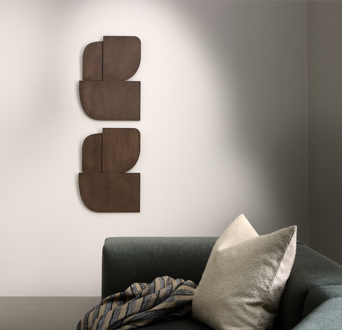 Minimalistiskt vardagsrumshörn med mörkgrön soffa, beige kudde och mönstrad filt; två vertikala geometriska väggpaneler i trä på en ljus vägg.
