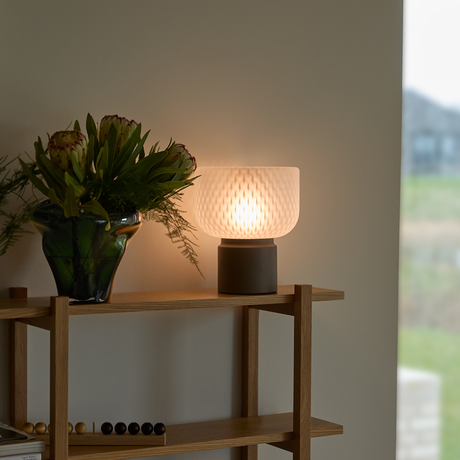 Tänd bordslampa i frostat glas på en trähylla bredvid en grön vas med blommor vid ett fönster.