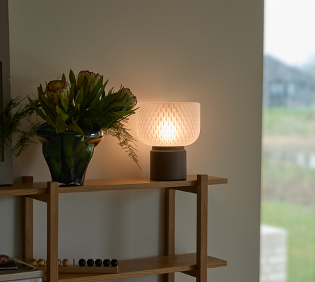 Bordslampa med mjukt sken bredvid grön glasvas med blommor på trähylla i vardagsrum med dagsljus från fönster.