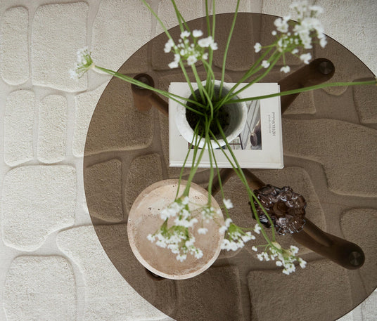 Rakt uppifrån av ett runt soffbord i glas med vitblommor i vas, en bok, en ljus stenskål och ett brunt glasobjekt på en beige, strukturerad matta.