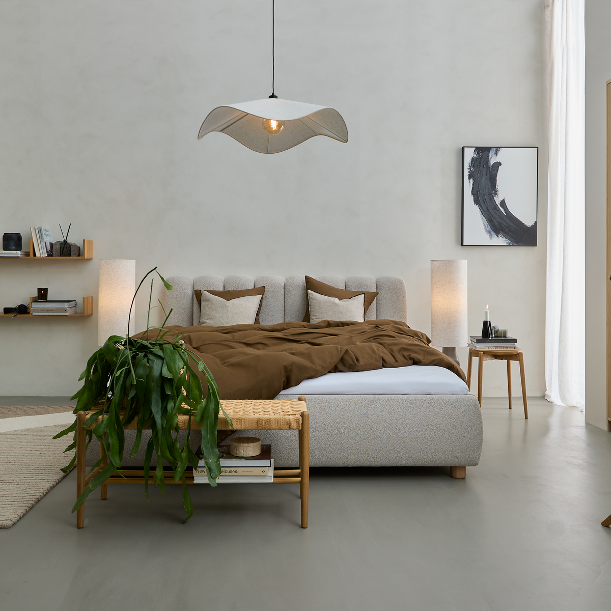Lugn, minimalistisk sovrumsscen med stoppad säng, brun bäddning, träbänk med hängande grön växt och vågformad pendellampa; mjukt dagsljus och abstrakt tavla på väggen.