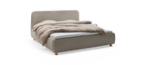 Stoppad dubbelsäng i ljusgrått tyg med beige täcke och runda träben, frilagd produktbild i sned vinkel._ps