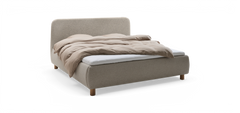 Uppklädd säng med rundad huvudgavel i ljus textil, beige täcke och synliga träben, fotad snett framifrån på neutral bakgrund.